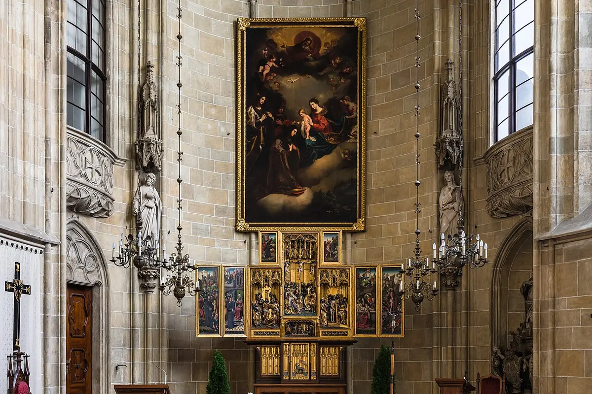 The chancel of the Deutschordenskirche in Vienna: a Gothic star vault, tall windows, a large altar painting above a gilded winged altarpiece and rows of wooden pews.